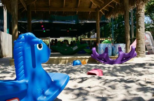 Colorful playground equipment, including a blue rocking horse, sits in sandy play area under a shaded structure.