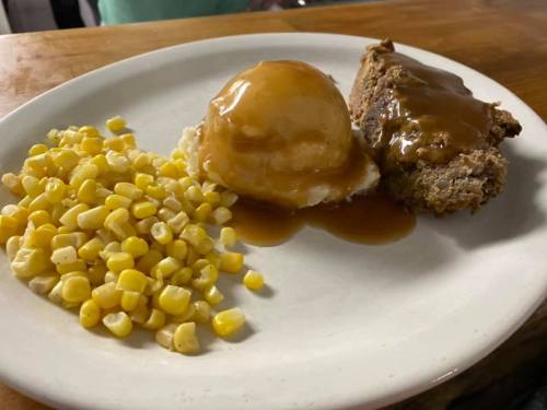 A plate with meatloaf topped with gravy, mashed potatoes with gravy, and a side of corn.