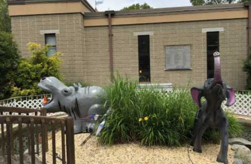 Two colorful animal sculptures, a hippo and an elephant, surrounded by greenery and gravel in front of a building.