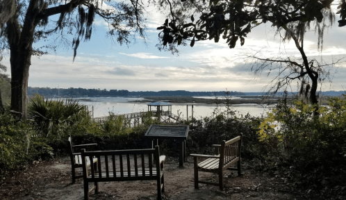 A serene view of a river with wooden benches and a sign, surrounded by trees and Spanish moss under a cloudy sky.