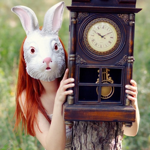 A person with a rabbit mask holds an antique clock, standing behind a tree in a green outdoor setting.