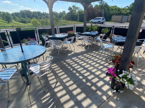Outdoor seating area with metal tables and chairs, shaded by a pergola, surrounded by flowers and greenery.