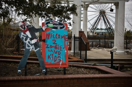 A graffiti-covered sign reading "Welcome 2 Zombie Land Kids" with a clown figure in front of an amusement park backdrop.