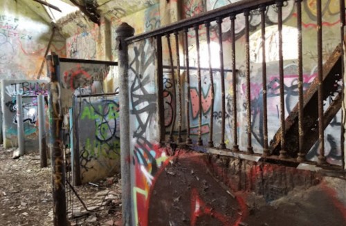 An abandoned room with graffiti-covered walls and a rusty staircase leading to a broken railing.