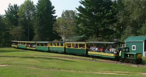 A green and yellow train with passengers travels along a scenic track surrounded by trees and grass.