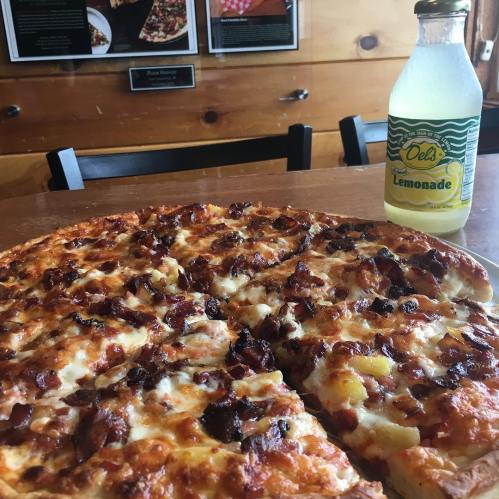 A close-up of a pizza topped with cheese and bacon, alongside a bottle of Del's Lemonade on a wooden table.