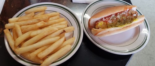 A plate with a hot dog topped with pickles and onions, alongside a serving of golden French fries.