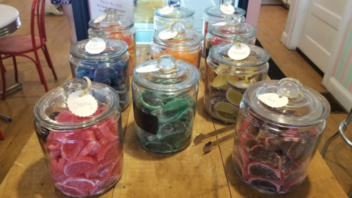 Colorful jars filled with various gummy candies, each labeled with different flavors, displayed on a wooden table.