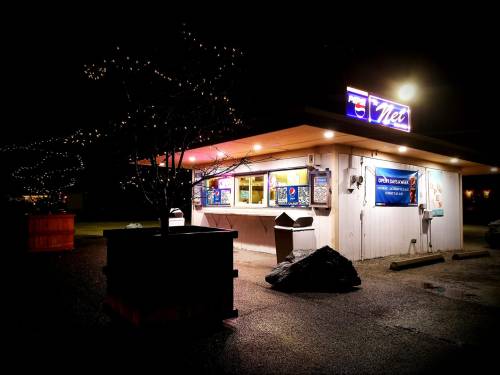A brightly lit food stand at night, featuring colorful signs and surrounded by trees and decorative lights.