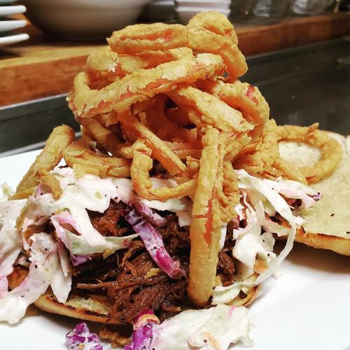 A hearty sandwich topped with crispy onion rings, shredded coleslaw, and tender pulled meat on a soft bun.
