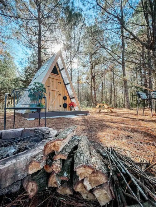 A cozy A-frame cabin surrounded by tall trees, with a firewood stack in the foreground and sunlight shining through.