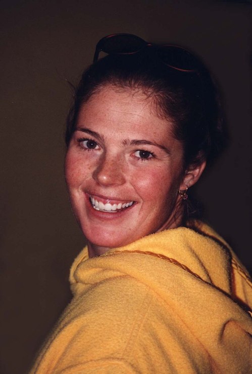 Smiling young woman in a yellow hoodie, with sunglasses on her head, looking over her shoulder.