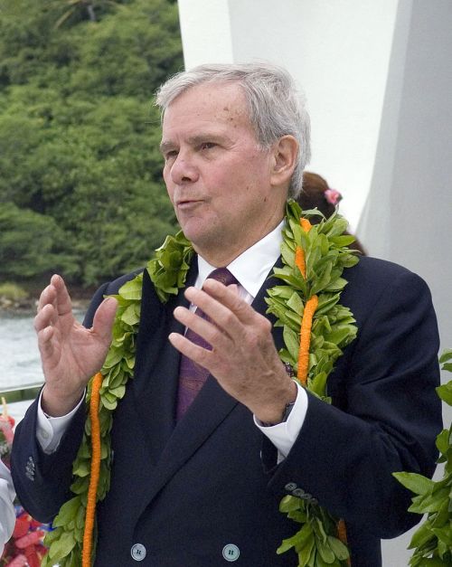 A man in a suit and floral lei gestures while speaking outdoors, surrounded by greenery and flowers.
