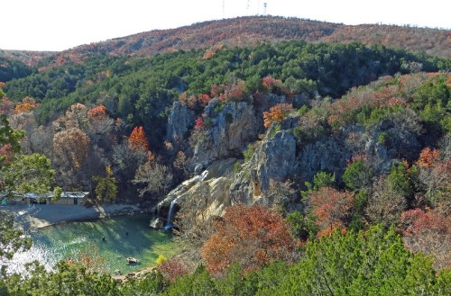 A scenic view of a waterfall cascading into a green pool, surrounded by autumn-colored trees and rocky hills.