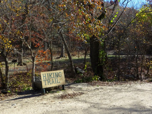 A wooden sign reading "Hiking Trail" stands beside a dirt path surrounded by trees in a wooded area.