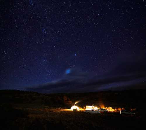 A starry night sky over a campsite with a glowing tent and parked vehicles in a remote landscape.