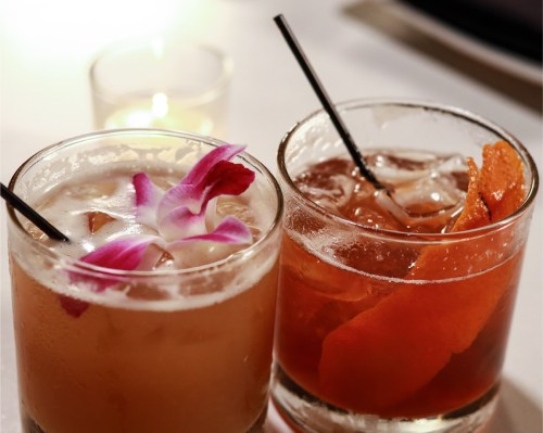 Two cocktails in glasses, one garnished with pink orchid petals and the other with orange peel, on a table with a candle.
