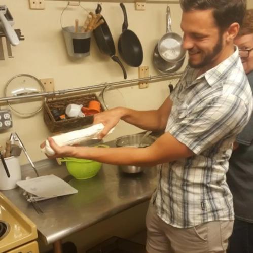 A man in a kitchen holds a rolled dough while another person stands nearby, surrounded by cooking utensils and ingredients.