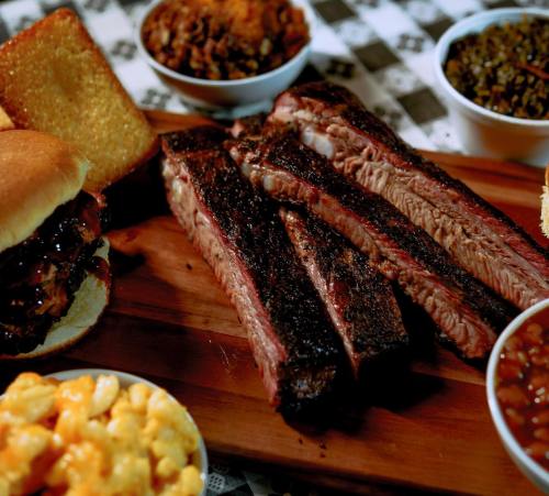 A wooden platter with sliced smoked brisket, cornbread, macaroni and cheese, baked beans, and collard greens.