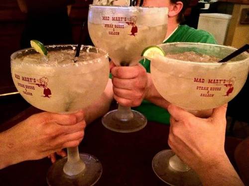 Three hands holding large margarita glasses with lime wedges, featuring the logo of Mad Mary's Steak House Saloon.