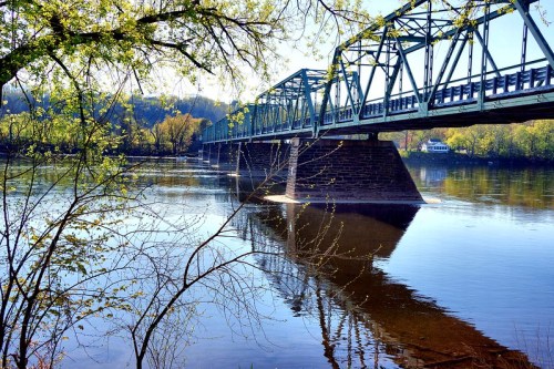 A green metal bridge spans a calm river, reflecting trees and blue sky in the water below.