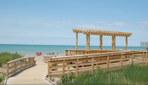A wooden walkway leads to a beach with colorful chairs and a calm blue lake under a clear sky.
