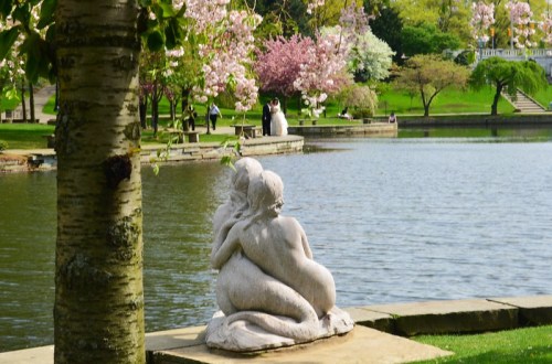 A serene park scene featuring a statue of two figures by a lake, surrounded by blooming trees and couples enjoying the view.