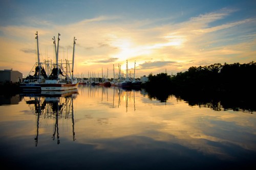 Sunset over a calm marina, with fishing boats reflecting on the water and a colorful sky in the background.