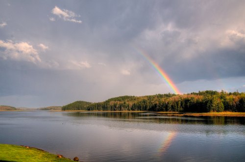 A serene landscape featuring a rainbow arching over a calm lake surrounded by lush green forests under a cloudy sky.