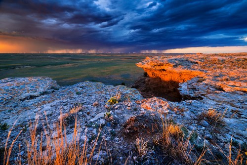 A dramatic landscape featuring rocky terrain, vibrant sunset colors, and dark storm clouds in the distance.