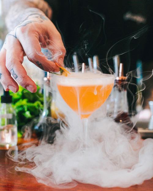 A hand garnishes a smoky orange cocktail in a glass, surrounded by mist and bar tools in the background.