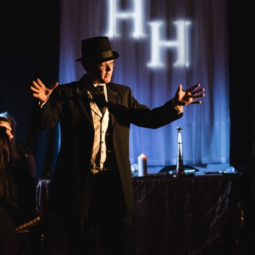 A performer in a black suit and hat gestures dramatically on stage, with a spotlight and candles in the background.