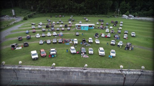 Aerial view of a grassy field filled with parked vehicles and small groups of people gathered around.