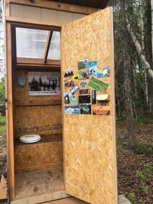 An open wooden outhouse with a toilet and various postcards and photos on the interior wall. Surrounded by trees.