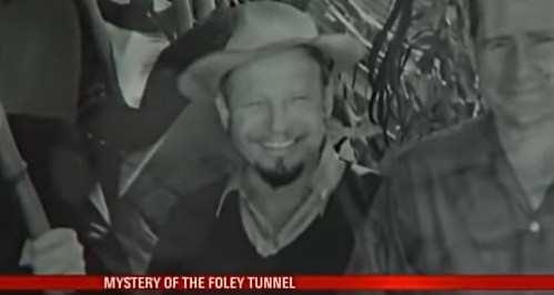 Black and white photo of a smiling man in a hat, surrounded by foliage and other individuals. Text at the bottom reads "Mystery of the Foley Tunnel."