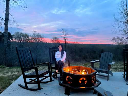 A person sits by a fire pit at sunset, surrounded by rocking chairs and a scenic view of trees and sky.