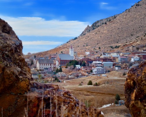 A scenic view of a small town nestled in a valley, surrounded by mountains and rocky terrain under a blue sky.