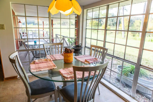 Bright dining area with a glass table, four chairs, and a view of a green outdoor space through large windows.