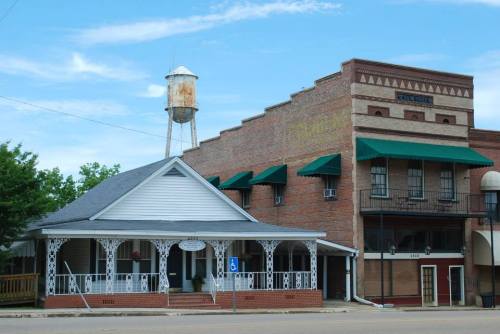 Historic buildings with a water tower in the background, featuring a white house with a porch and a brick structure.