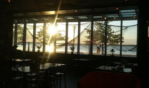 Sunset view through large windows in a cozy restaurant, with trees and water visible outside.
