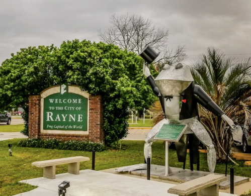 A large metal frog in a top hat stands next to a welcome sign for Rayne, the "Frog Capital of the World."