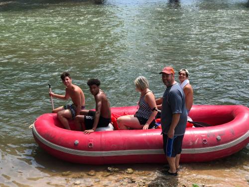 A group of five people in a red inflatable boat on a river, enjoying a sunny day outdoors.