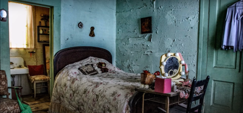 A vintage bedroom with a floral bedspread, a mirror, and rustic decor, featuring peeling blue walls and soft lighting.