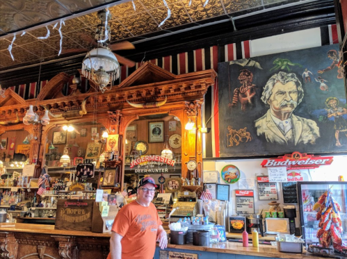 A man stands in a vintage bar with ornate woodwork, colorful decor, and a large painting of Mark Twain on the wall.
