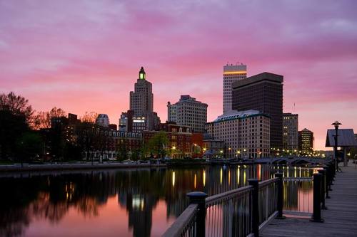 City skyline at sunset, reflecting in a river with a colorful sky and trees along the waterfront.