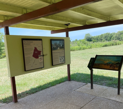 Information signs under a shelter in a grassy area, with trees and blue sky in the background.