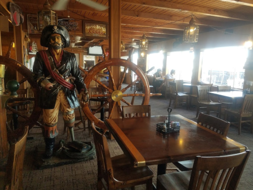 A pirate statue stands near a wooden table in a cozy restaurant with nautical decor and large windows.