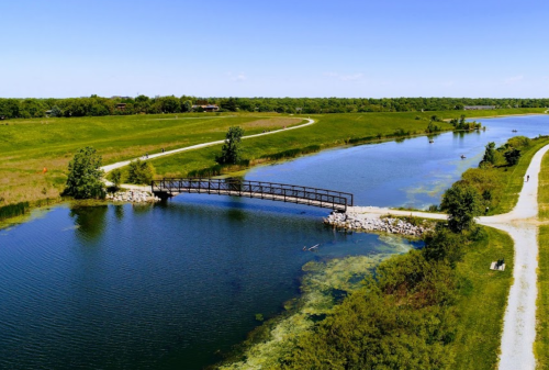 A serene landscape featuring a bridge over a calm lake, surrounded by green fields and walking paths.