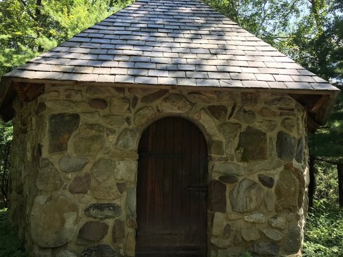 A small stone building with a sloped roof and wooden door, surrounded by trees in a natural setting.