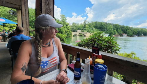 A woman with a braid sits at a lakeside restaurant, looking out at the water and greenery, with drinks on the table.
